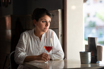 Glass of rose wine is held by a dark-haired girl by a bar window.