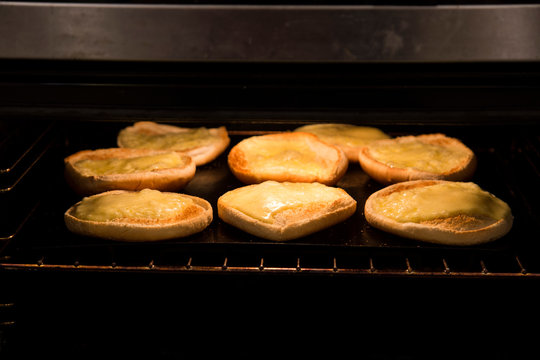 Slices of mild cheddar cheese under the grill for home made family burger night. Cheese is yellow, bread rolls are white and oven is dark with stainless steel edges. Hamburgers are a family favourite.