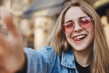 Girl chilling and having fun, taking selfie while on vacation in foreign city. Charming happy european female with fair hair, pulling hand towards camera, smiling and sticking out tongue