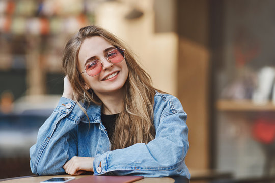 Girl likes to eat a lot. Tender charming fair woman in denim jacket, touching and playing with hair, wearing trendy glasses while leaning on table and smiling at camera, sitting in chinese restaurant
