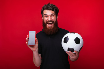 Joyful young and bearded man holds a soccer ball and a smartphone on red background.