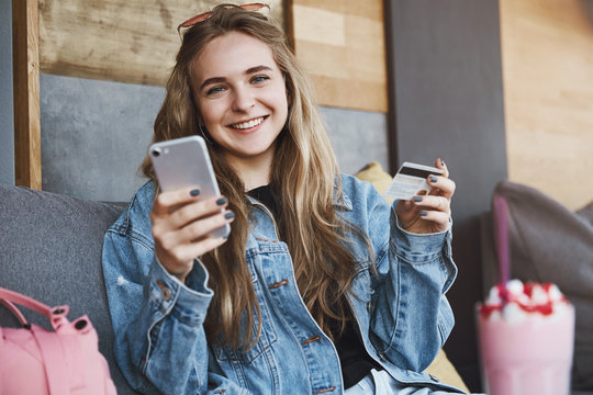Friends sitting in cute cafe, drinking and talking while attractive blonde student in trendy jacket checking bank account via smartphone, typing credit card number in app, smiling joyfully