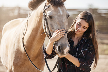 Obraz premium Animals, friendship and hobby concept. Pretty young woman in rural area smiling, gently petting horse nose, vet taking care of domestic pets on countryside farm, teaching children become jokey