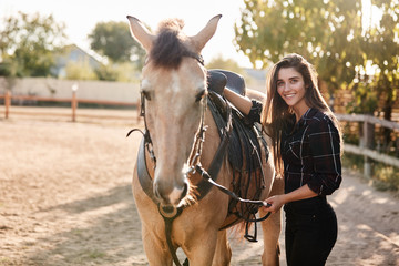 Fototapeta premium Girl prepare her horse for ride on racetrack. Cheerful female jockey fasten saddle, grooming cute animal and smiling joyfully camera, enjoy fresh air, taking care rancho, farm pets