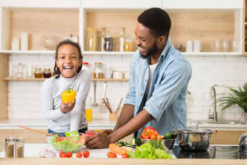 Cheerful afro girl holding pepper while helping father cooking dinner