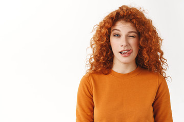 Playful cute stylish redhead curly-haired female student enjoy life, lead active lifestyle, showing tongue and wink joyfully, stay bright side, fool around cheerfully standing white background