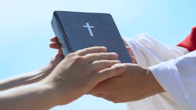 Male hands taking Bible from priest in robe, spread of Christian doctrine