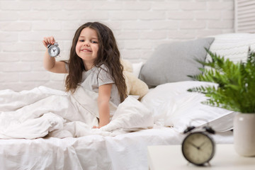 cute smiling little child girl in pyjamas with clock in bed. good morning
