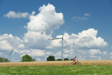 nice, active woman, riding her electric mountain bike between wheat fields and wind wheewls of a wind farm on the Schwaebische Alb near the city of Aalen, BadenWuerttemberg, Germany