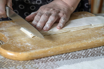 Senior woman cuts the dough on wooden board during homemade noodle or pasta production in her home kitchen. Closeup, selective focus