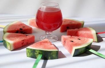 Close-up fresh watermelon juice in a glass with a straw and sliced ​​watermelon with colored sticks around the glass on a white background with stripes of shadows.