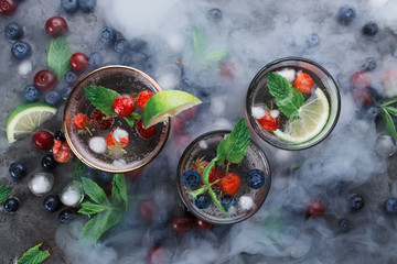Flatlay of three cups with iced soda water on dark background with mint, herbs, berries and ice and smoke, selective focus