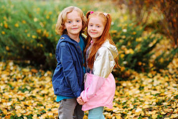 two children - a small boy with blond curly hair and a girl with red hair hugging holding hands against the background of autumn yellow leaves and Park. The concept of autumn, love, friendship.