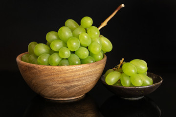 Lot of whole fresh green grape in big wooden bowl in dark ceramic bowl isolated on black glass