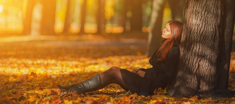 Beautiful woman in black coat and leather boots sitting under tree in autumn park with fallen leaves. Bright young long haired brunette girl relax in park with red yellow maple leaves in fall season.