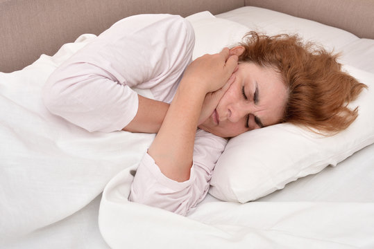 Mature woman with toothache lying in bed and holding her cheek