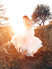 Woman in white dress and fashionable hat walking on field in sunset. Golden hour. Fashion.