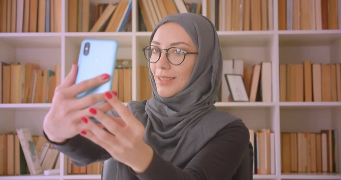 Closeup portrait of young attractive muslim businesswoman in glasses and hijab taking selfies on phone sitting in front of laptop in library indoors