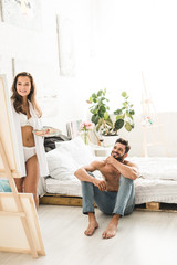 full length view of girl drawing with brush and man sitting on floor near bed and smiling while looking at painting