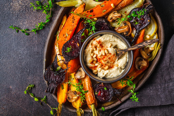 Baked vegetables with hummus in a dark dish, top view. - obrazy, fototapety, plakaty