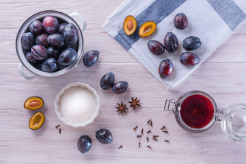 An open glass jar with homemade plum jam, fresh ripe plums, folded towel, badyan seeds and dry carnation grains on a wooden table. Flat lay, top view