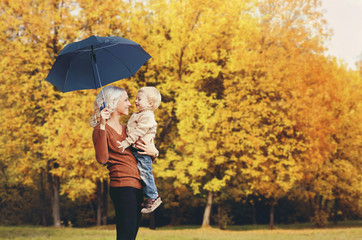 Happy smiling mother holding child with umbrella having fun together over autumn trees background