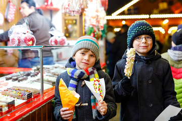 Obraz premium Two little kid boys, cute siblings eating bananas covered with chocolate, marshmellows and colorful sprinkles near sweet stand with gingerbread and nuts. Happy children on Christmas market in Germany.