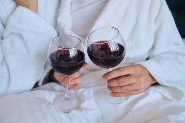 Close up man and woman in white bathrobes holding glasses of red wine in hands lying in bed in modern hotel