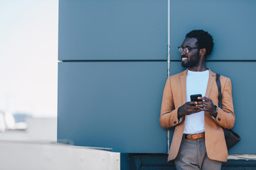 smiling african american businessman standing on rooftop and looking away while holding smartphone