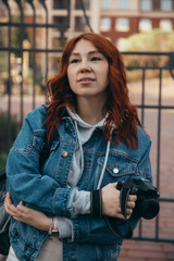 Beautiful red-haired girl posing with a camera on the street near a brick building on a summer evening