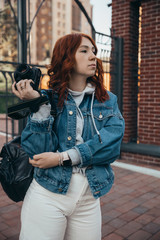 Beautiful red-haired girl posing with a camera on the street near a brick building on a summer evening