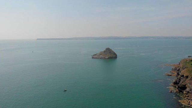 An aerial forward footage of a rocky island rock (Thatcher Point) with speeding boat in the background along rocky cliff and turquoise water under a majestic blue sky