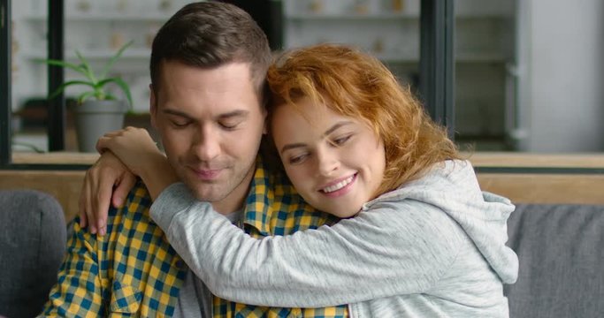 Young pretty woman is hugging man, doing online shopping, holding credit card, they are sitting on grey sofa at home, Caucasian. 4K, shot on RED camera.