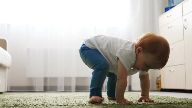 Baby girl falling down during learning to walk on floor in bright apartment.