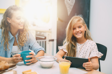 portrait of cute blond little girl using tablet while having breakfast with her mother at home