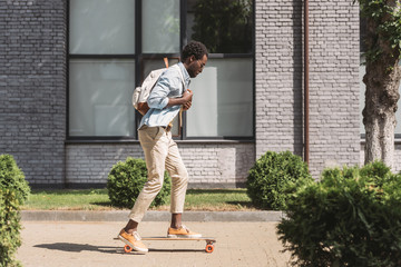 stylish african american man with backpack riding longboard on sunny street