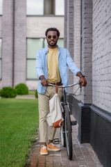 Fototapeta na wymiar handsome, stylish african american man in sunglasses looking at camera while standing with bicycle near brick wall