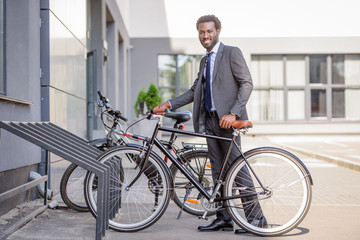 Fototapeta premium handsome african american businessman smiling at camera while taking bike from parking