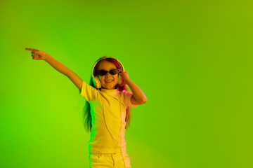 Beautiful female half-length portrait isolated on green backgroud in neon light. Young emotional teen girl. Human emotions, facial expression concept. Dancing in sunglasses and pointing up.