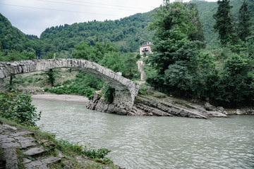 Fototapeta na wymiar Medieval bridge over a mountain river, trees along the banks of the river, power lines and the road hidden behind the foliage of trees. Cloudy day with clouds in the sky.
