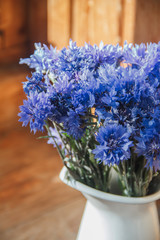Flowers of a cornflower in a white iron pitcher. The pitcher stands on a wooden shelf. A bouquet of flowers in a jug. Blue flowers in a vase.