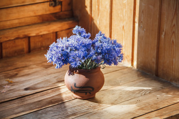 Flowers of cornflower in white earthenware. The vase is on a wooden shelf. A bouquet of flowers in a vase of clay. Blue flowers in a vase.