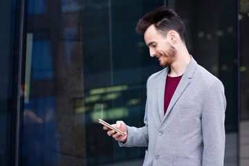 Businessman is looking at screen of cell phone and smiling. Side view. Smart casual style, formal dressed guy with a beard standing outdoor office and typing or texting a message or sms on smartphone.