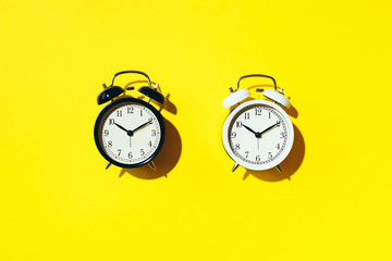 Black alarm clock and white one with hard shadow on yellow background. Top view