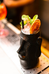 Cocktail in black glass with dried orange and mint on the bar, blurred background.