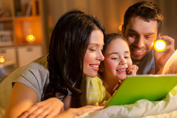 people and family concept - happy mother, father and little daughter reading book with torch light in bed at night at home