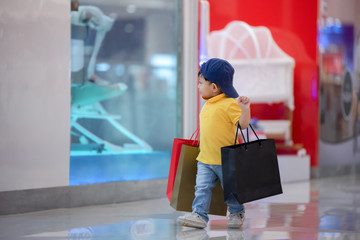 Kid shopping. Asian little boy in yellow shirt and jeans enjoyment of shop. Walk in the shopping mall and holding heavy shopping paper bags.