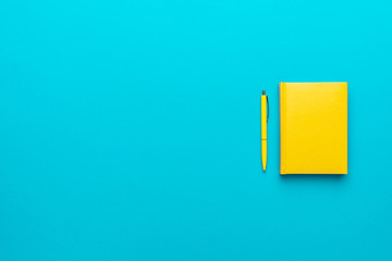 Top view photo of closed yellow notebook and ball-point pen over turquoise blue background with...