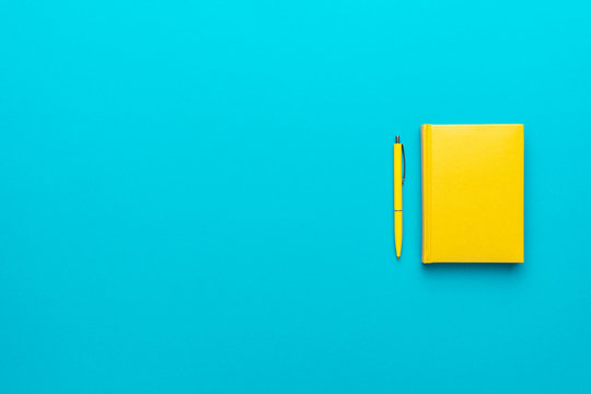Top view photo of closed yellow notebook and ball-point pen over turquoise blue background with copy space. Minimalist flat lay image of closed diary and yellow pen as back to school background.
