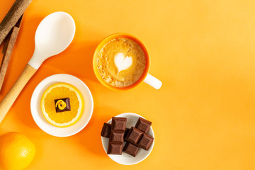 Chocolate pieces over orange slice, chocolate bars, full orange, latte coffee, cinnamon sticks and white cooking spoon over orange background with copy space.